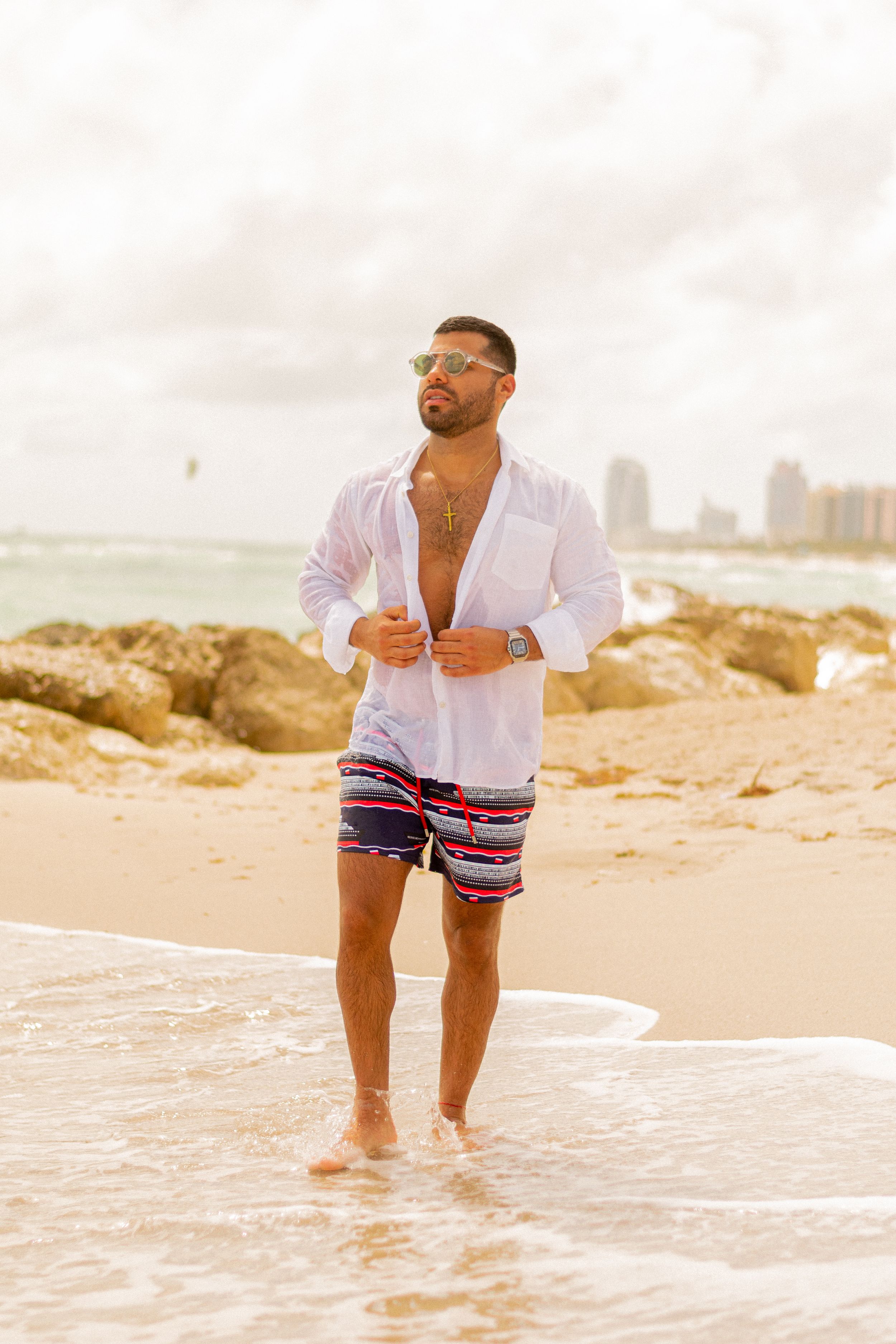 Person in white shirt and patterned swim shorts standing on beach shore at sunset.