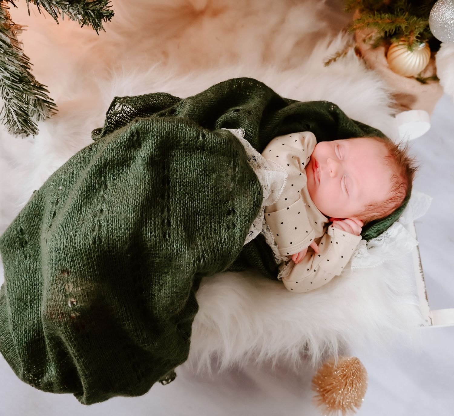 A sleeping baby wrapped in a green blanket rests on white fur under Christmas decorations.