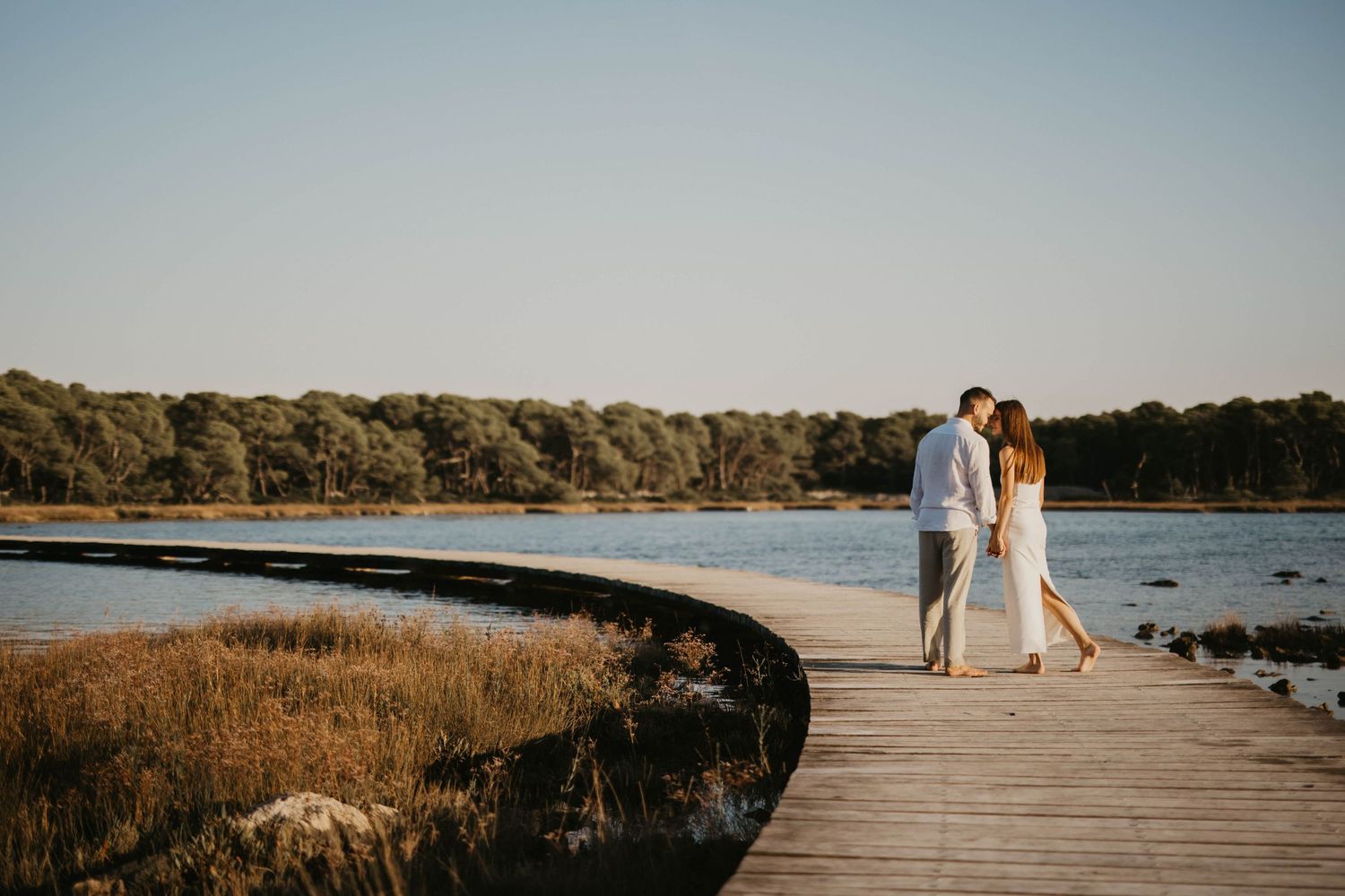 Couple walking along wooden boardwalk by waterfront during sunset.