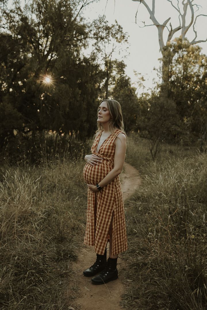 Pregnant woman in a plaid dress stands on a dirt path at sunset in Majura Park, with sunlight filtering through trees in the background.