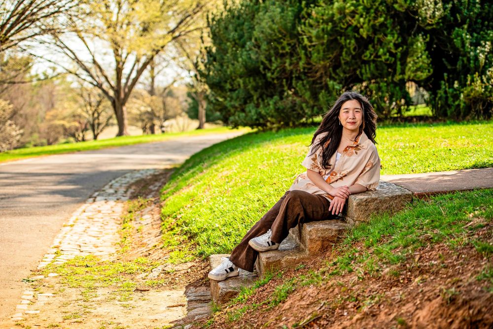 Woman sitting on stone steps in a sunny park with green grass and tree-lined path in the background.