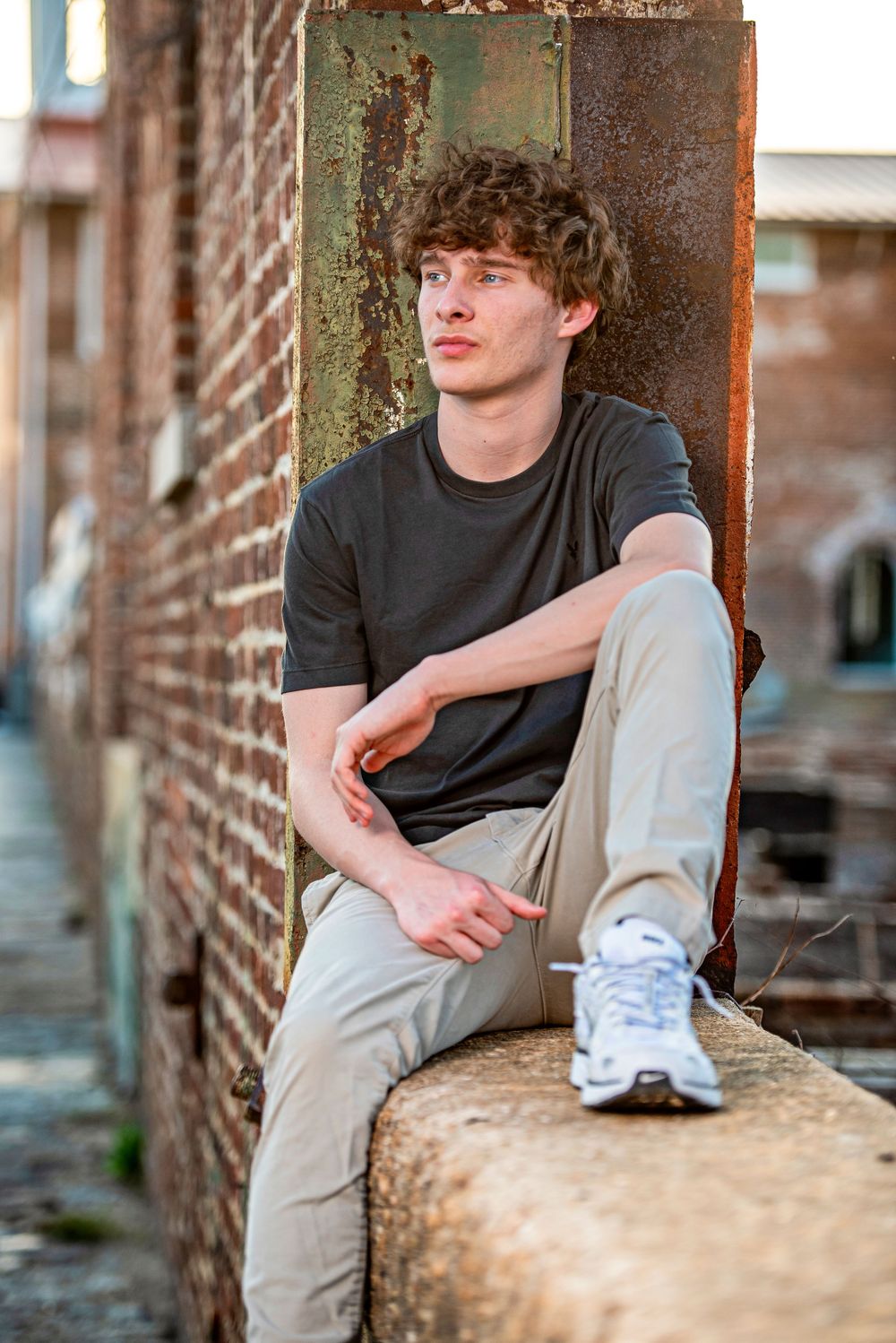 Young man with curly hair sitting on ledge against rustic brick wall, wearing gray t-shirt and khaki pants.