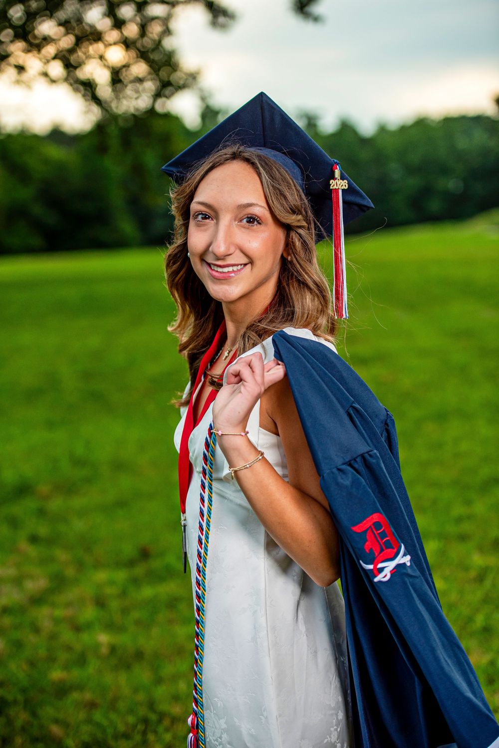 Smiling graduate poses outdoors in navy cap and gown with red honor cords on a lush green field.