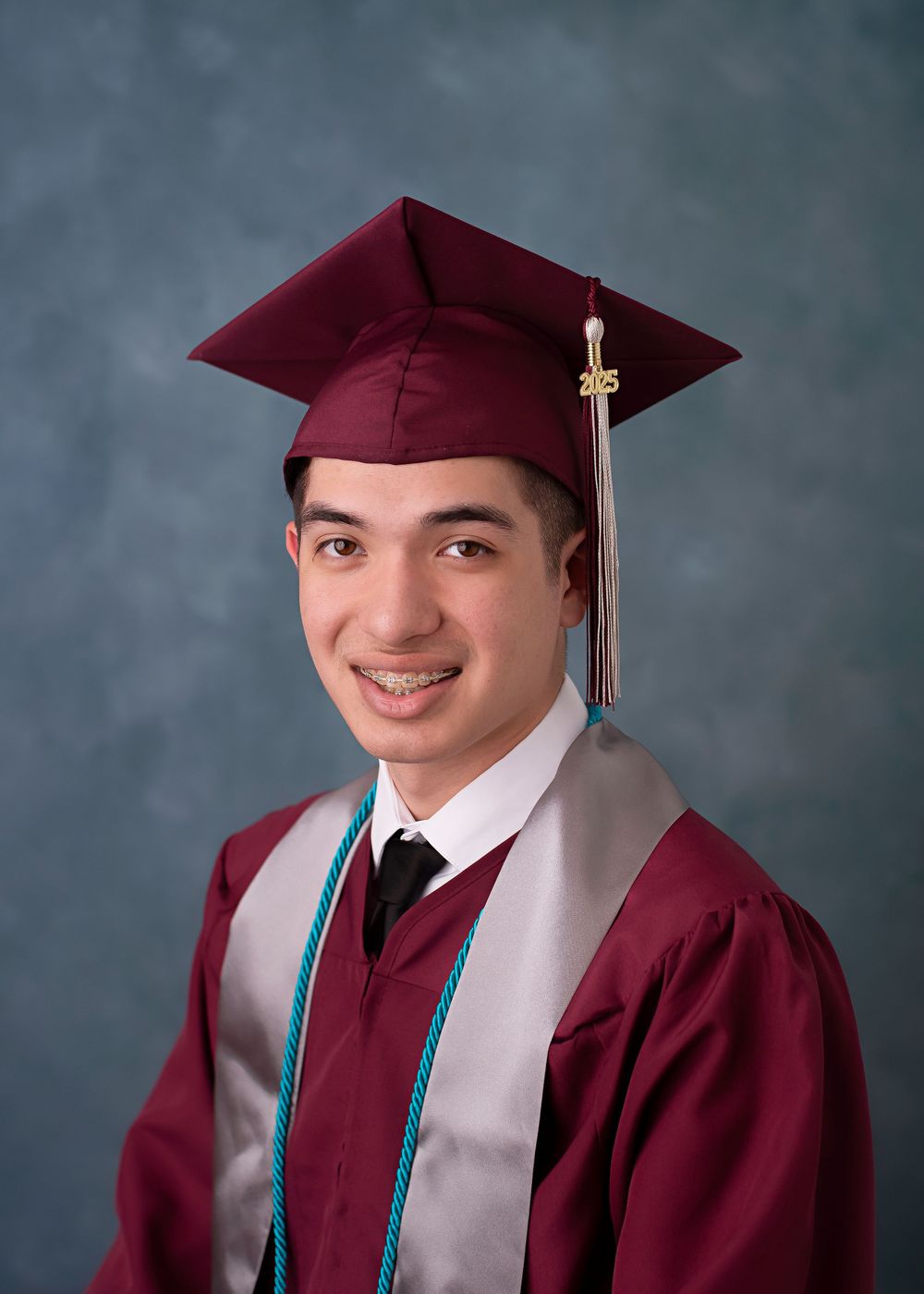 Student posed in maroon graduation cap and gown with silver stole against gray backdrop at commencement ceremony.