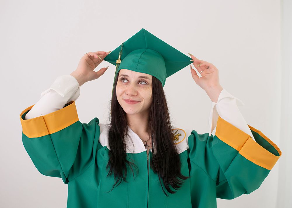 A graduate student adjusts their green and gold graduation cap and gown while smiling against a white background.