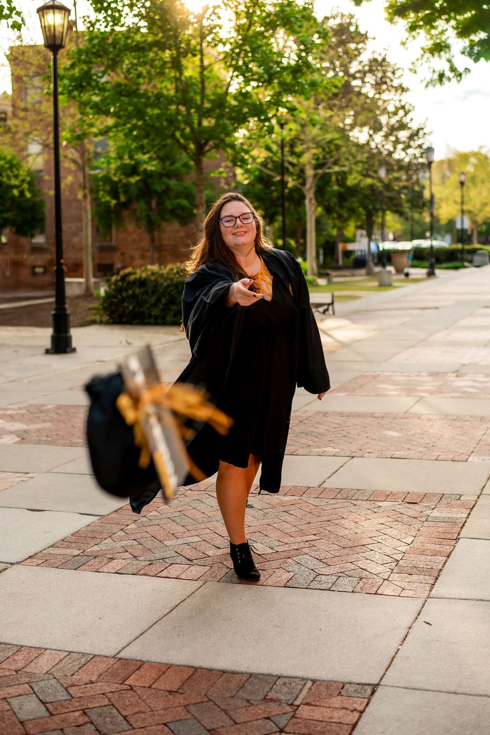 Graduate wearing black cap and gown walks joyfully down brick sidewalk near lamp post on sunny university campus.