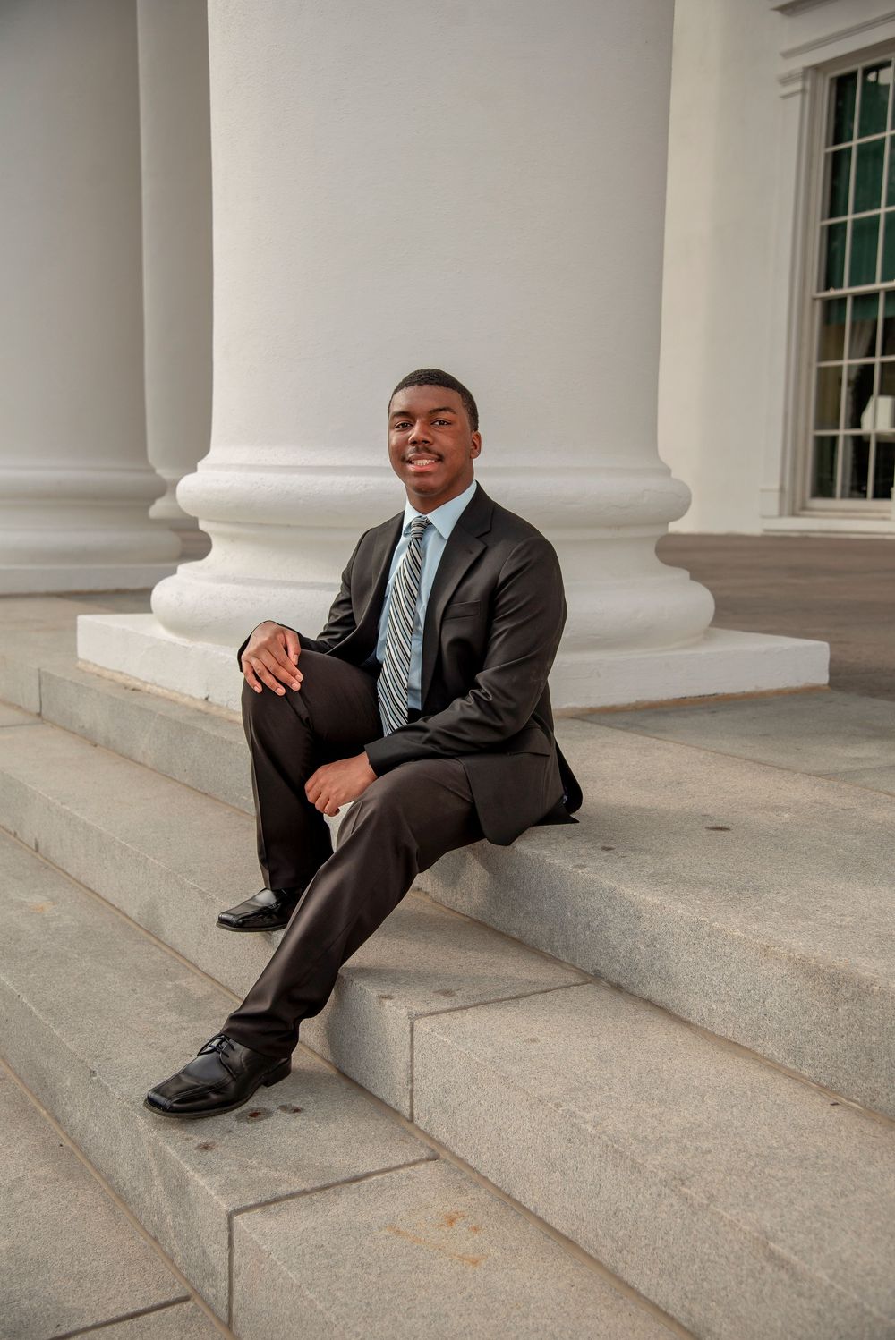 A professional in a dark suit and blue tie sits casually on stone steps with white columns in the background.