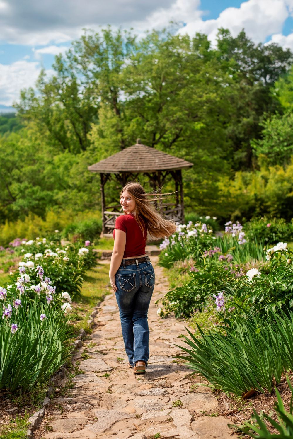 A stone pathway leads through a blooming garden with irises and a wooden gazebo in the background.