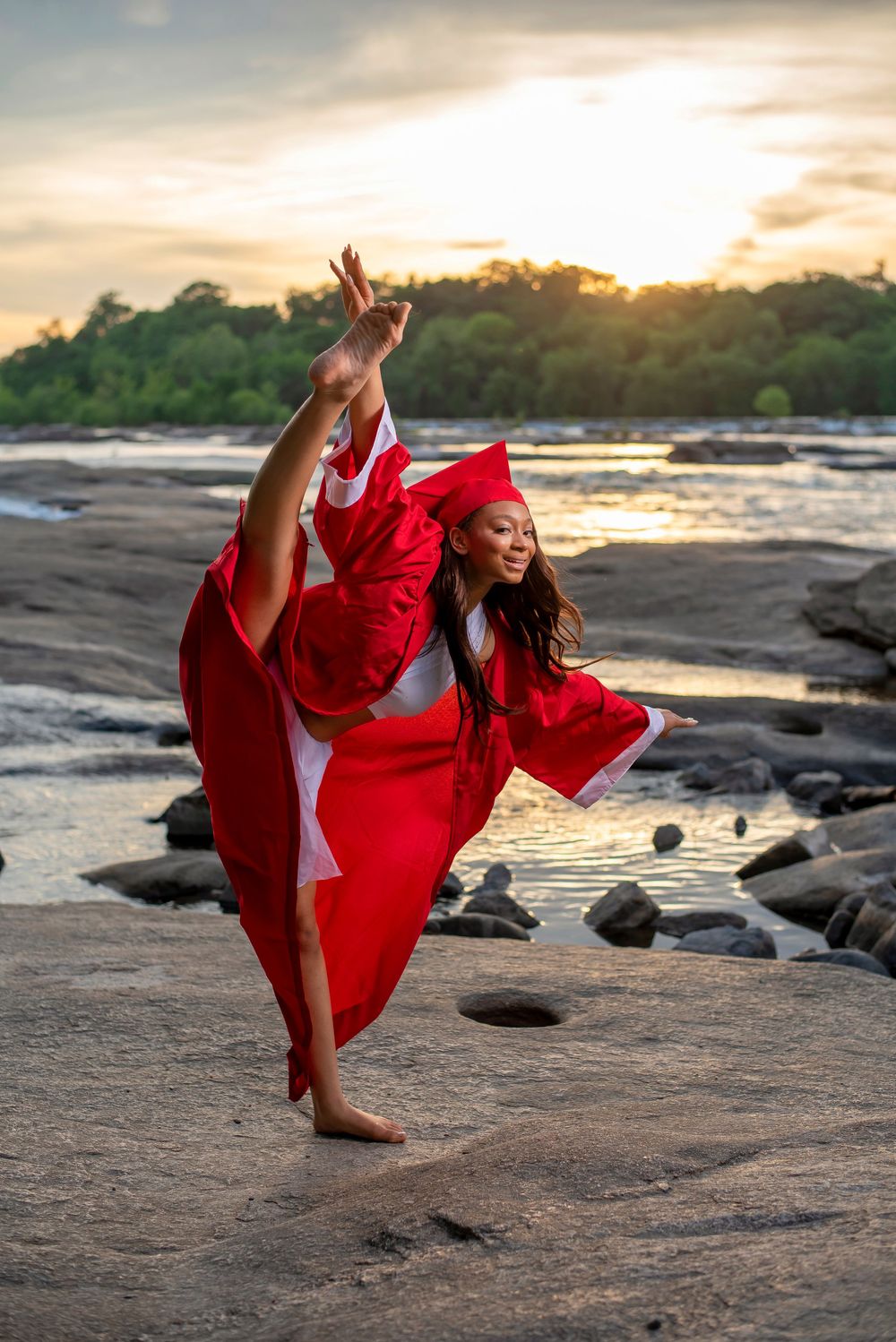 Dancer in flowing red dress performs graceful pose on rocky beach at sunset.
