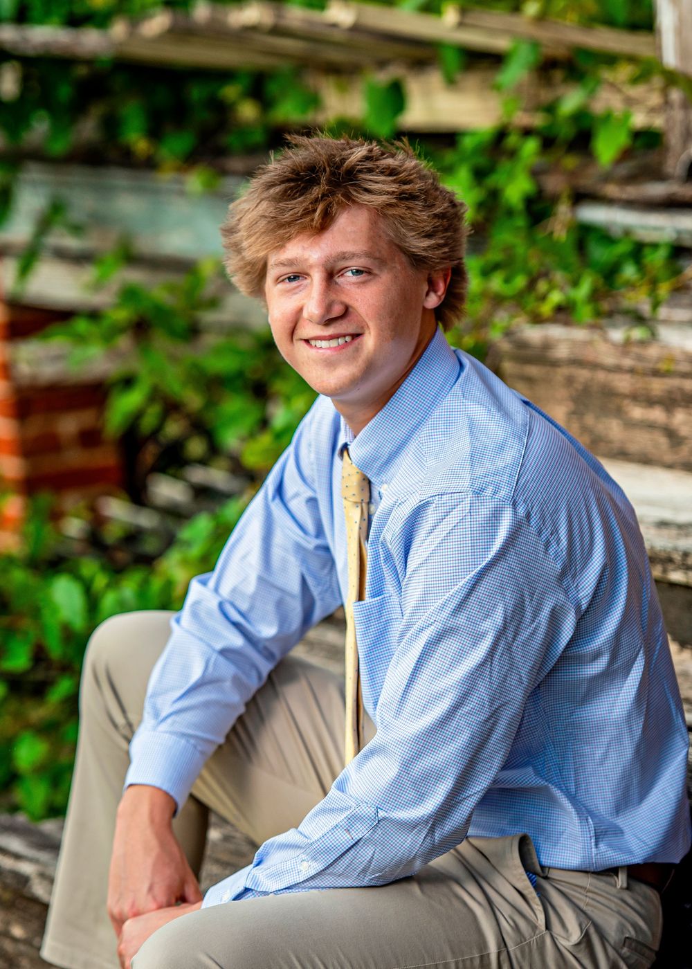 Smiling young man in light blue dress shirt and gold tie seated outdoors with green ivy background.