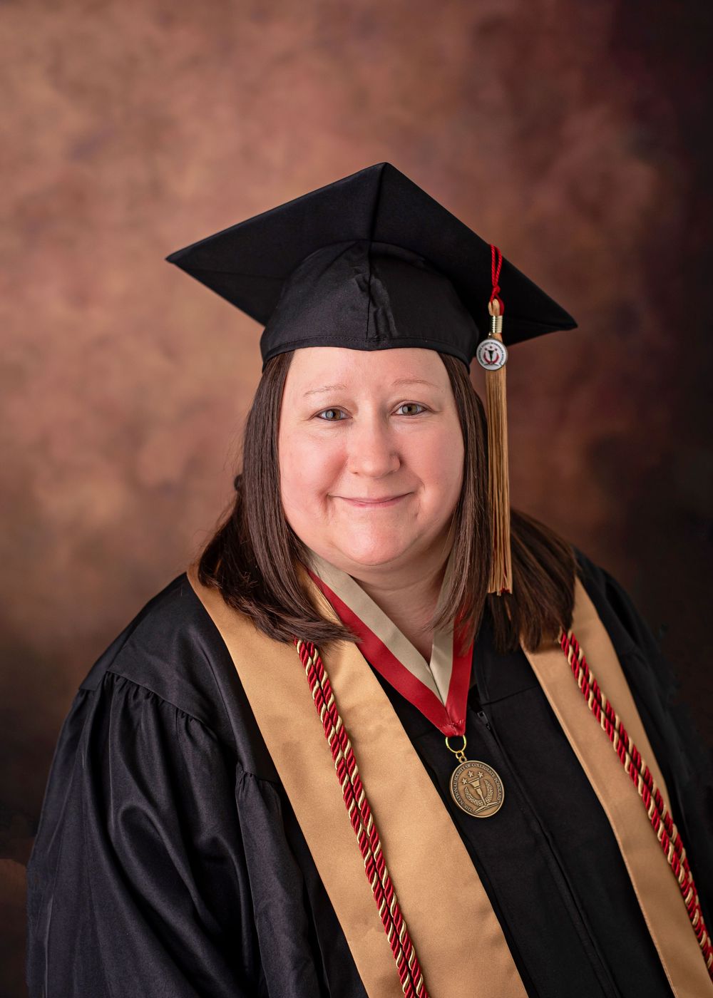 Woman in black graduation cap and gown with gold sash and medal smiling for formal graduation portrait.