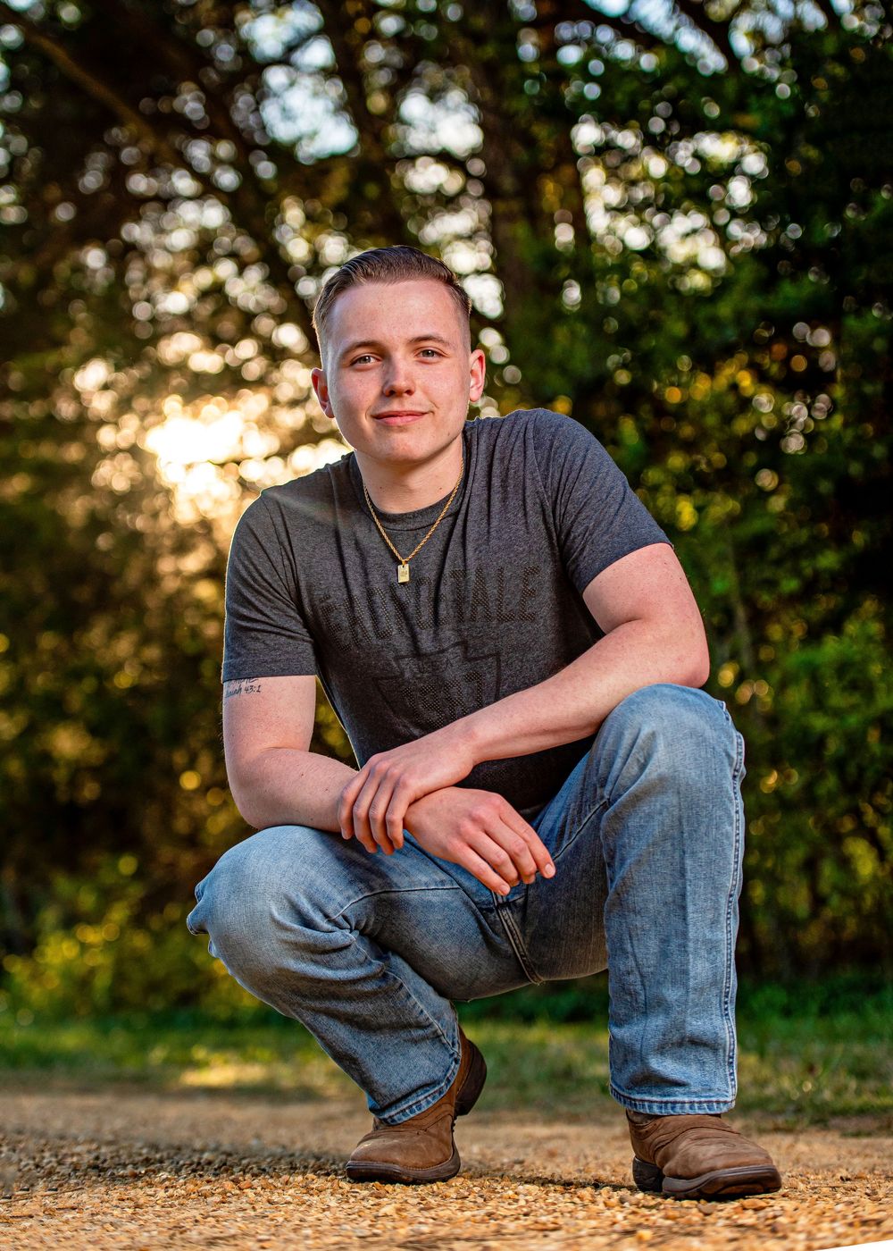 Young man in gray t-shirt and jeans crouching on a dirt path surrounded by lush green trees at golden hour.