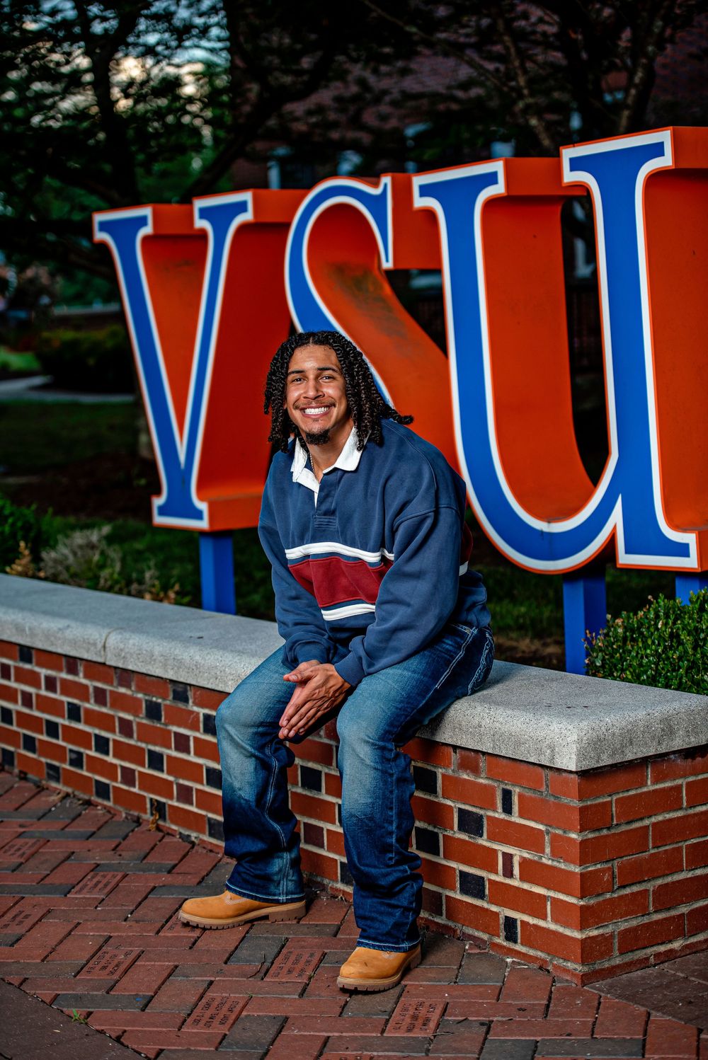 Smiling student sits on brick wall in front of VSU university sign on campus.