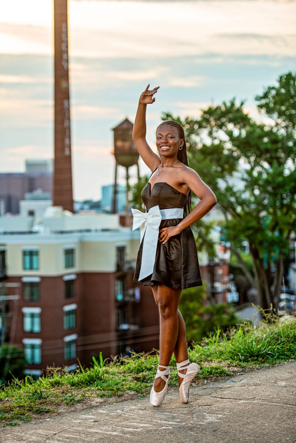 A Black ballerina in a dark dress with white bow poses en pointe outdoors with city buildings and water tower behind her.