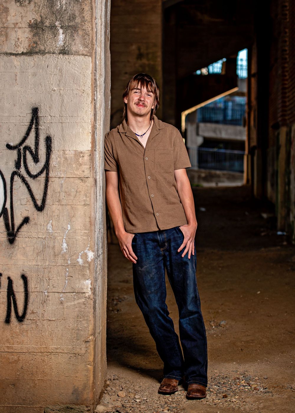 Young man in brown shirt and jeans leaning against graffiti-covered concrete pillar in urban setting.