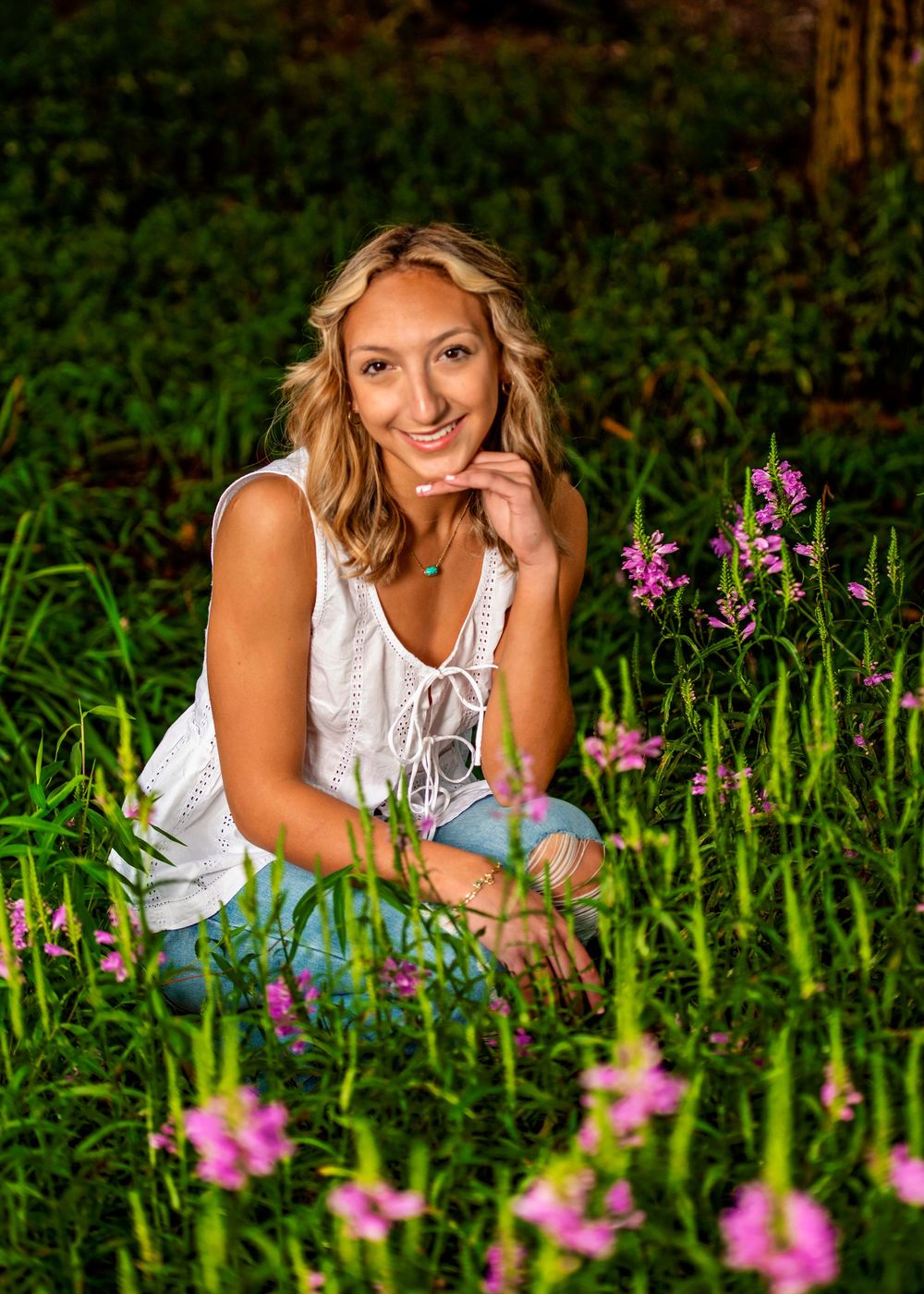 A woman in a white sleeveless top poses gracefully among pink wildflowers in a lush outdoor setting.