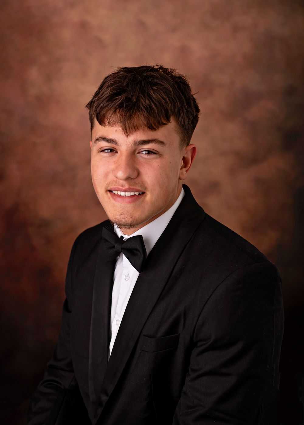 Young man smiling in a black tuxedo with bow tie, posing for a formal portrait photo.