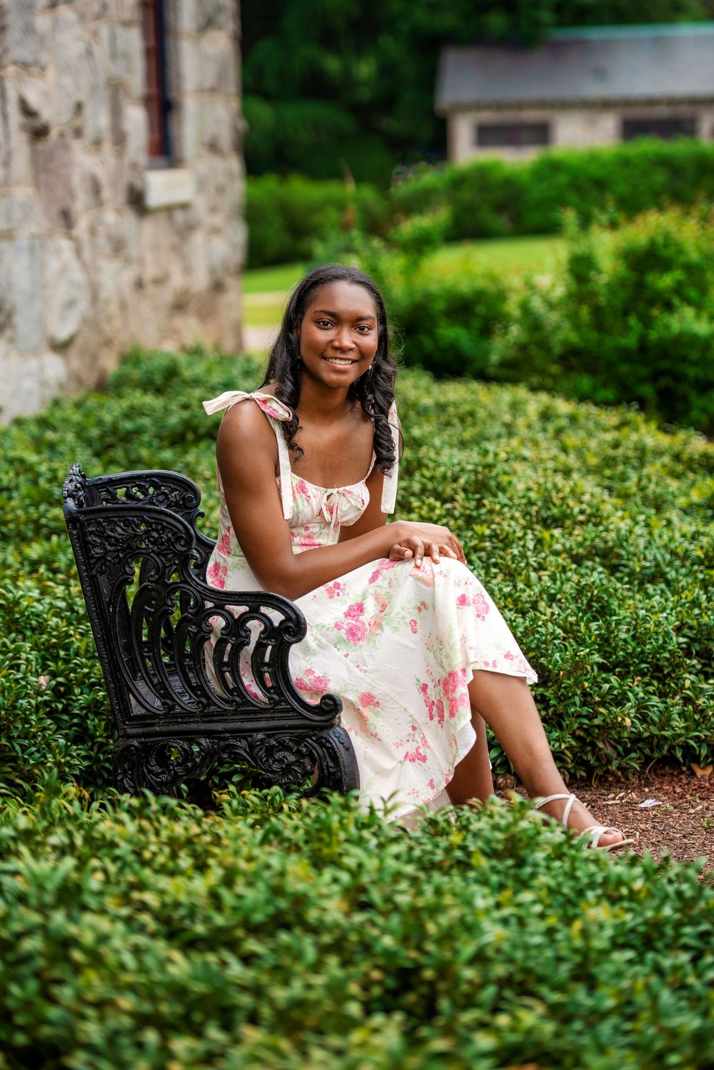 A person wearing a floral summer dress sits on an ornate black metal bench surrounded by manicured garden hedges.