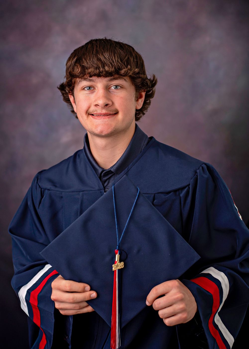 Young graduate smiling in navy blue cap and gown holding diploma at formal graduation portrait session.