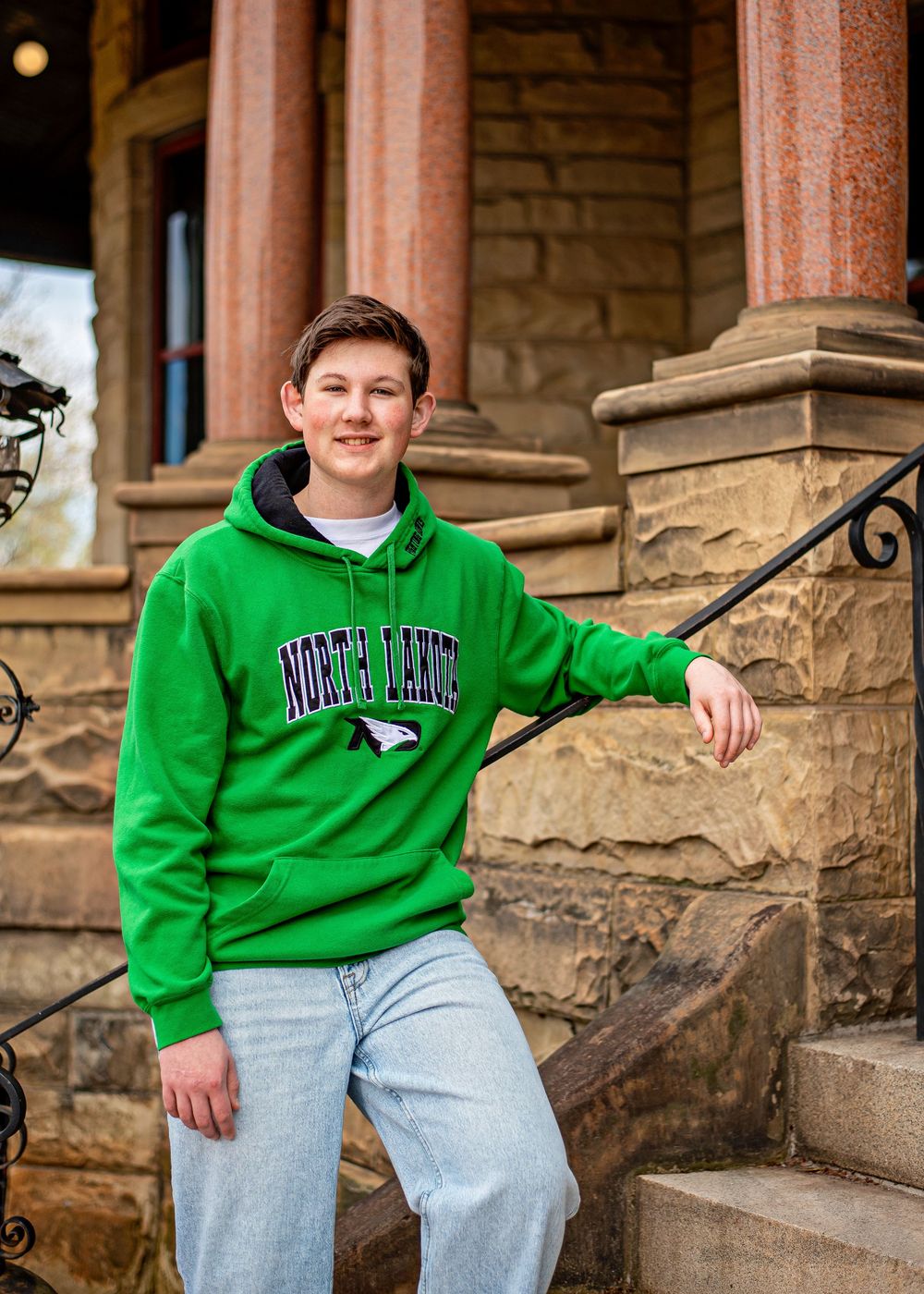 Young man in green North Dakota hoodie sitting on stone building steps, smiling for a portrait photo.