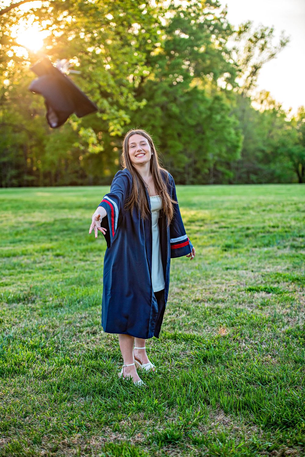 Smiling graduate in navy blue cap and gown tossing mortarboard in sunny green park at golden hour.