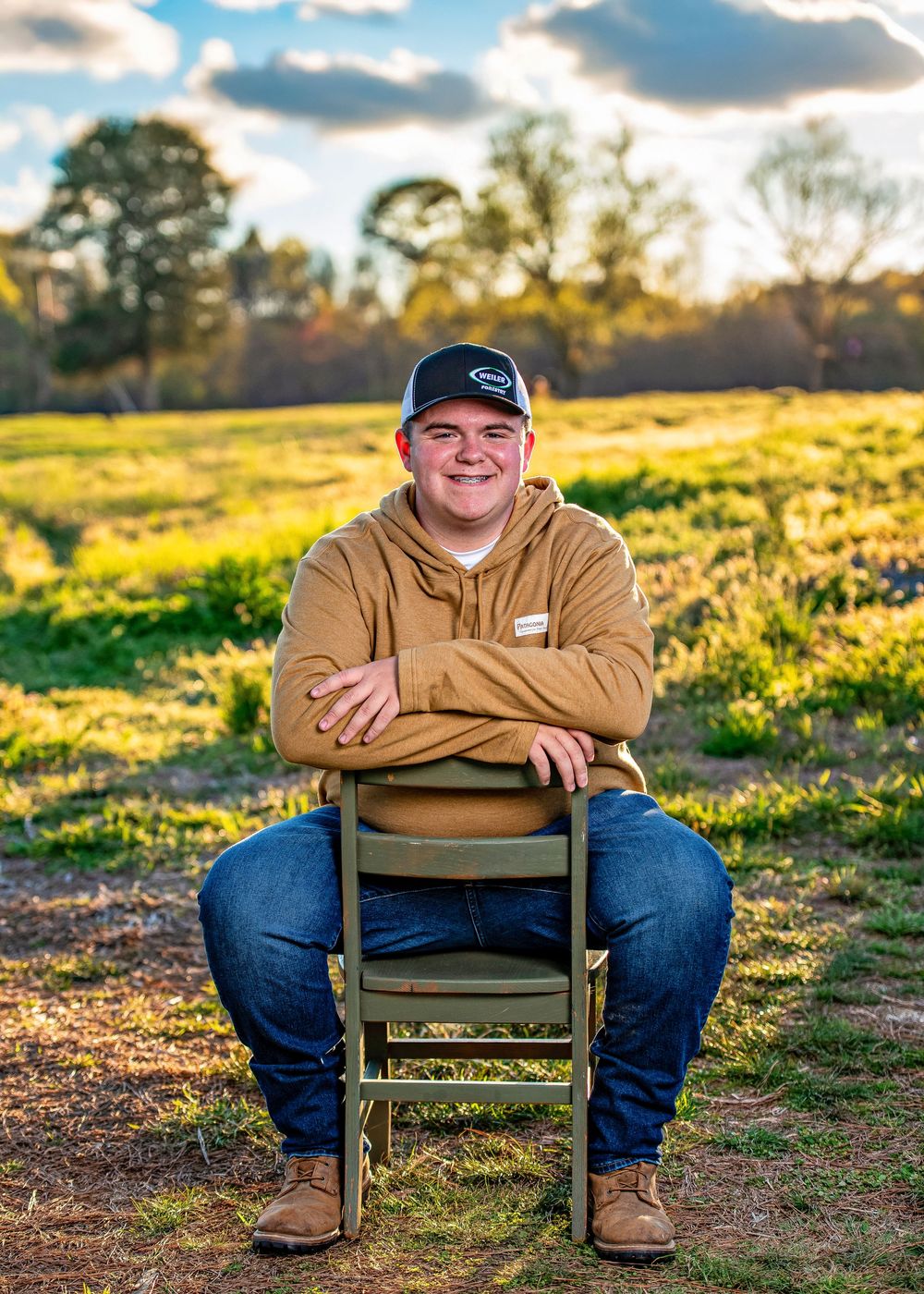 Young man in tan hoodie and cap sits cross-armed on chair in sunny green field for outdoor portrait.