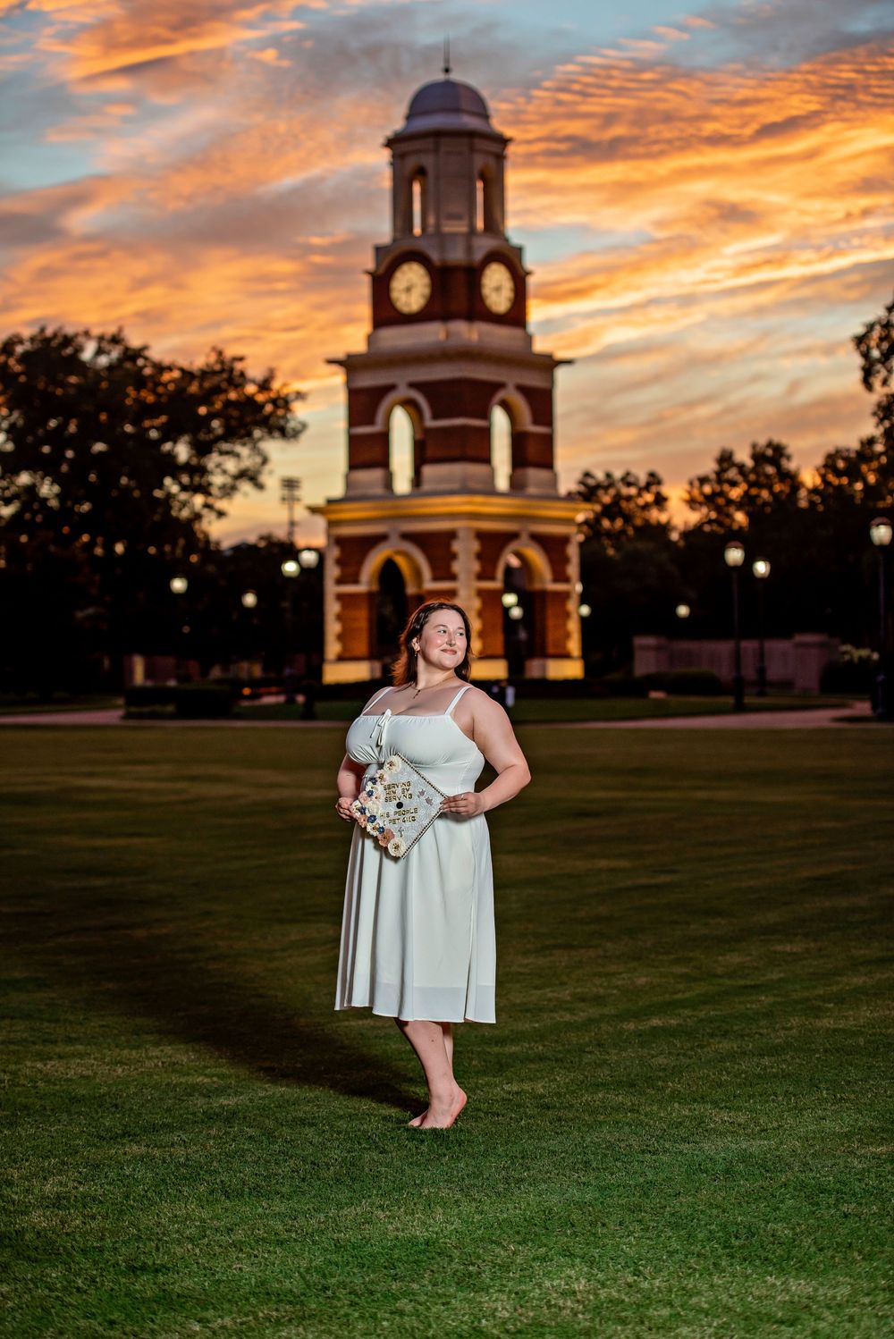 Woman in mint dress poses before a brick clock tower at sunset with vibrant orange sky.