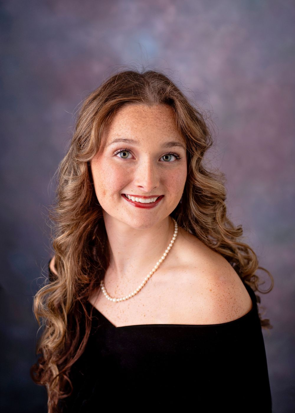 Young woman with curly auburn hair, freckles, pearl necklace, and black off-shoulder top smiling in a portrait photo.