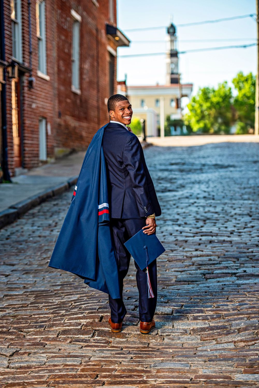 Smiling graduate in navy cap and gown walks cobblestone street, glancing back at camera near historic brick buildings.