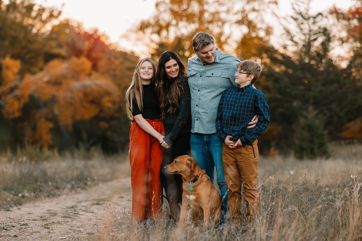 A family and their brown dog pose together in a field during golden hour with autumn colors.