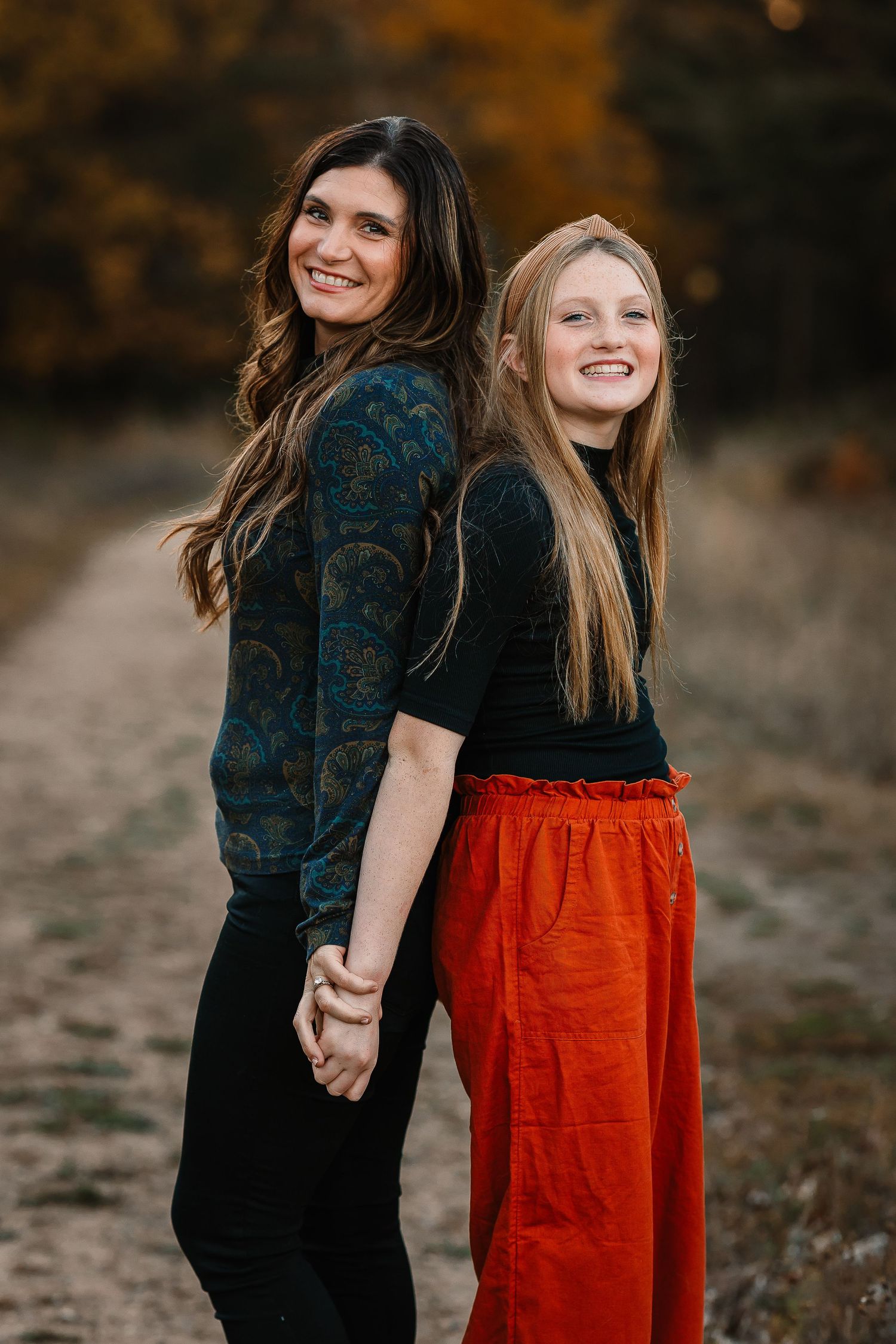 Two people stand back-to-back on a dirt path during autumn wearing coordinated outfits.