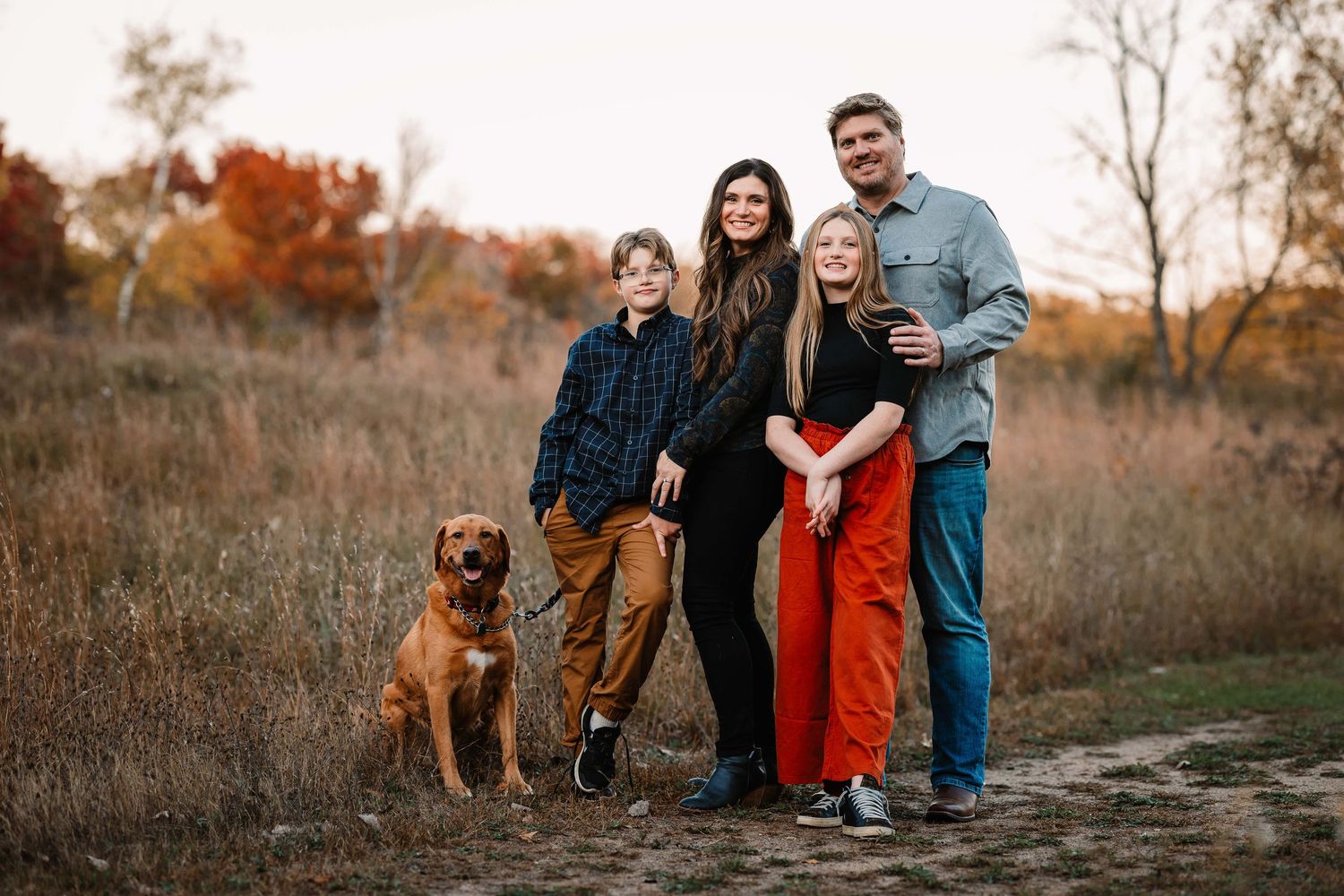 A family poses with their brown dog on a trail during autumn with fall foliage in the background.