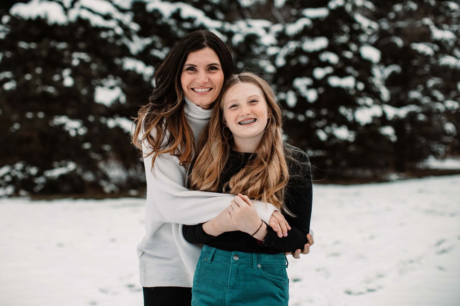 Two people embrace while smiling against a snowy evergreen backdrop.