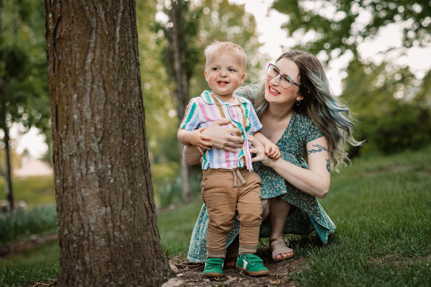 Parent and child sit under trees in park during playful outdoor photoshoot with natural lighting.