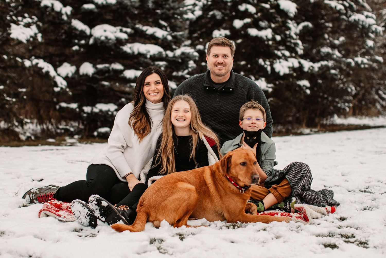 A family with their brown dog sits together on snowy ground in front of evergreen trees.