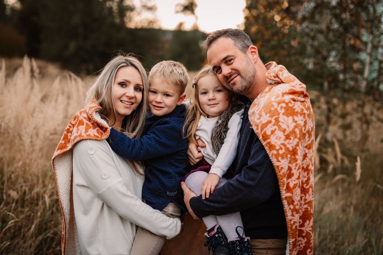 A family embraces outdoors during a fall photo session while wearing warm toned clothing against a natural backdrop.
