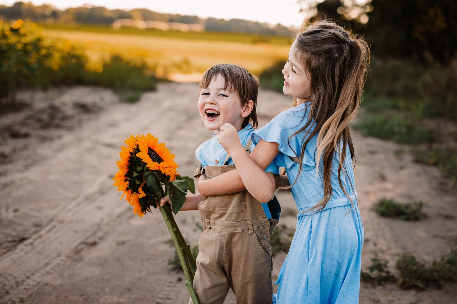Two children share a joyful moment with sunflowers on a dirt path at golden hour.