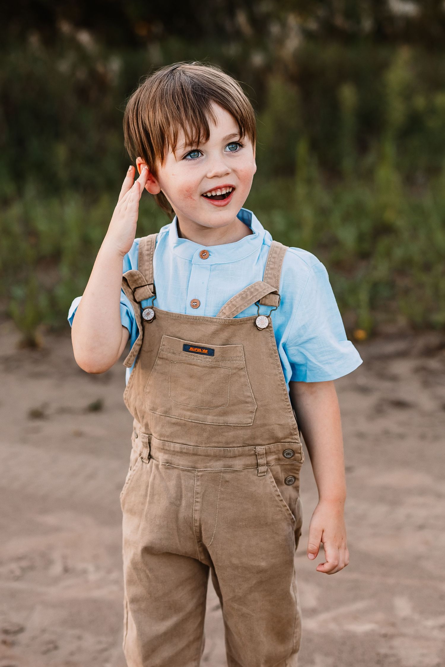 Young child wearing brown overalls and light blue shirt stands outdoors with a playful expression.