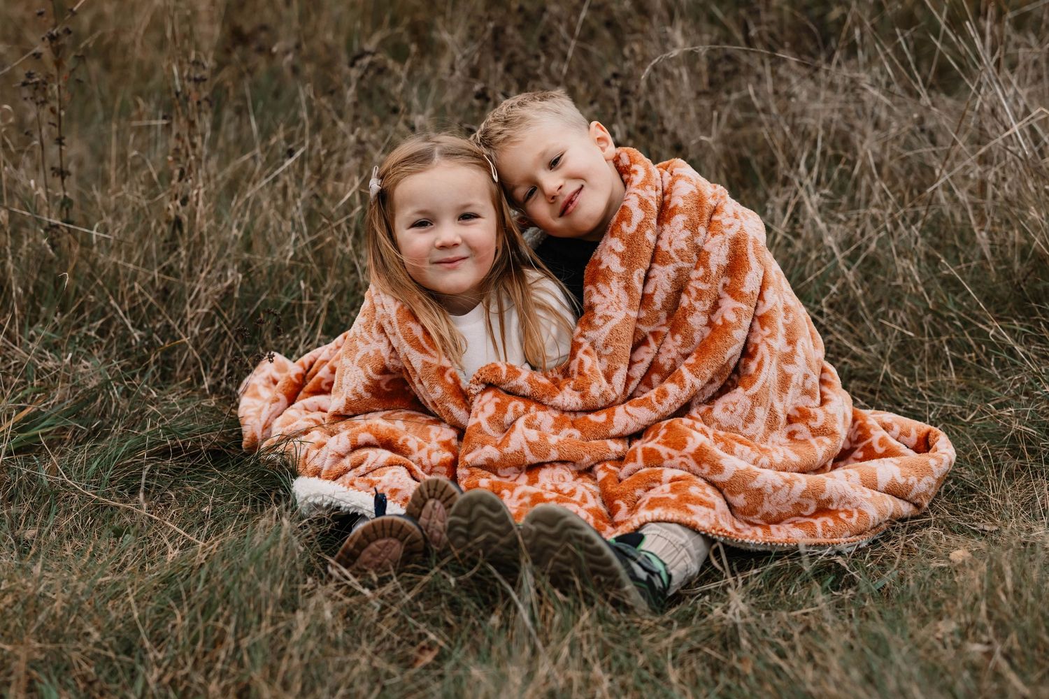 Two children snuggle under orange patterned blanket in autumn field.