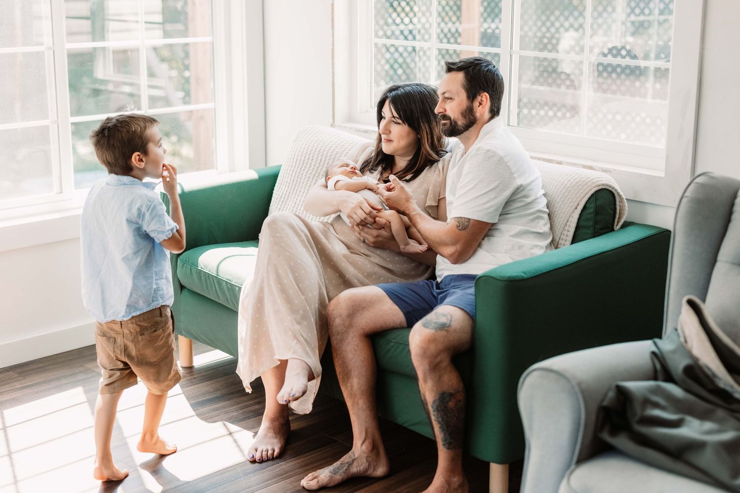 Family relaxes together on green couch while sharing tablet device in bright sunlit living room.