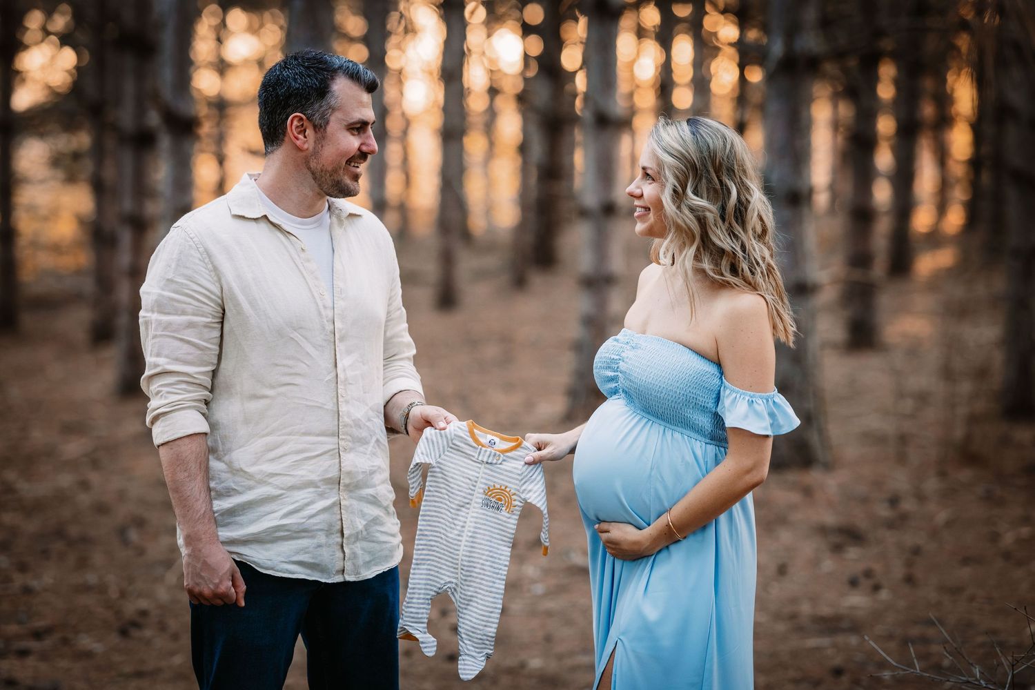 A couple holds baby clothes during a maternity photoshoot in a forest at sunset.