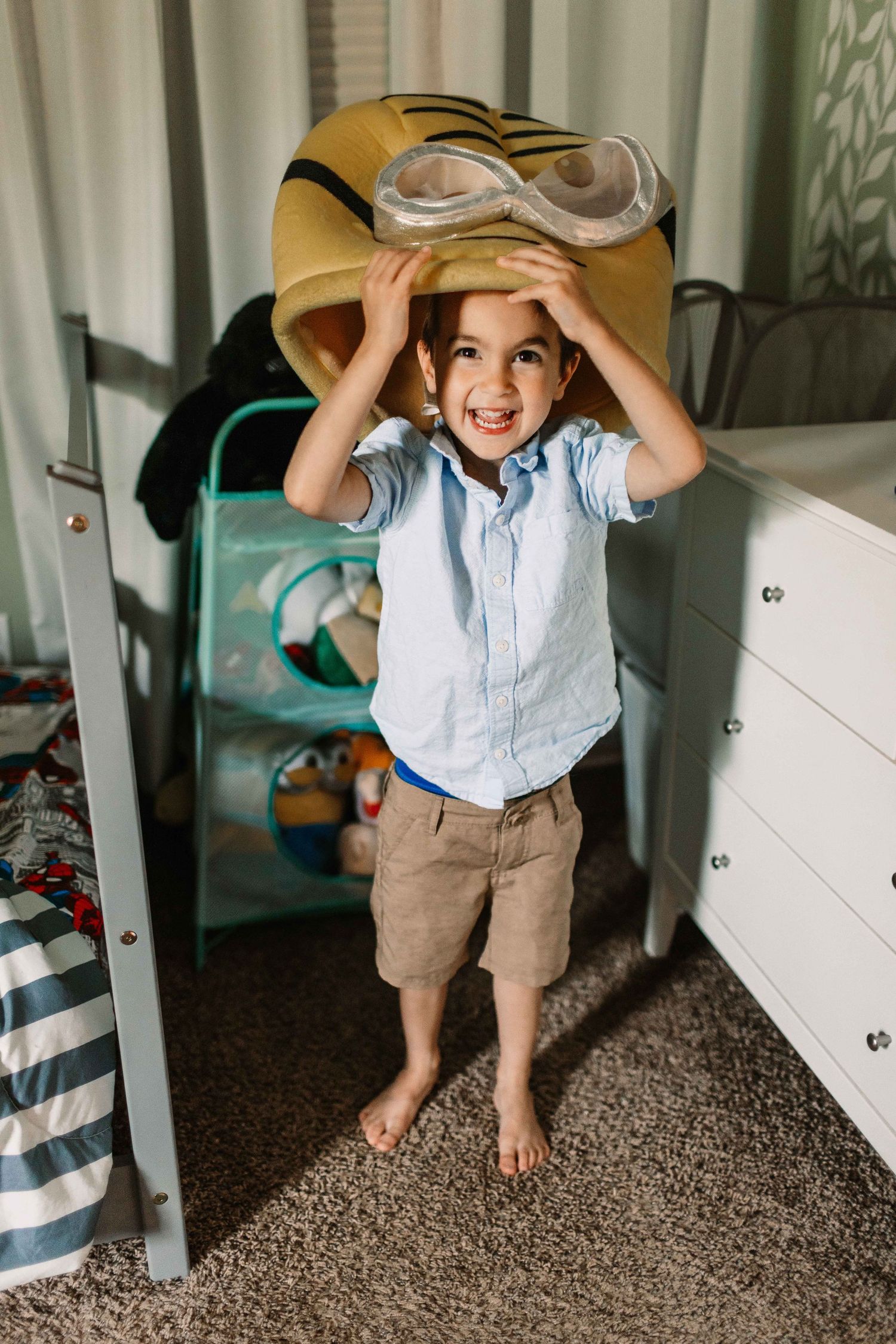 Small child in blue shirt and brown shorts plays peek-a-boo with a beige hat in bedroom corner.