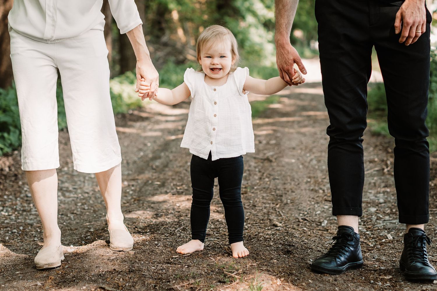 Small child takes first steps between two people on a forest path.