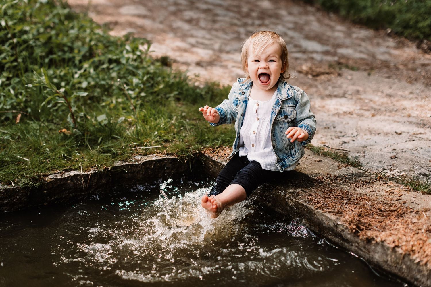 Excited toddler splashes in puddle on dirt path creating joyful water spray.