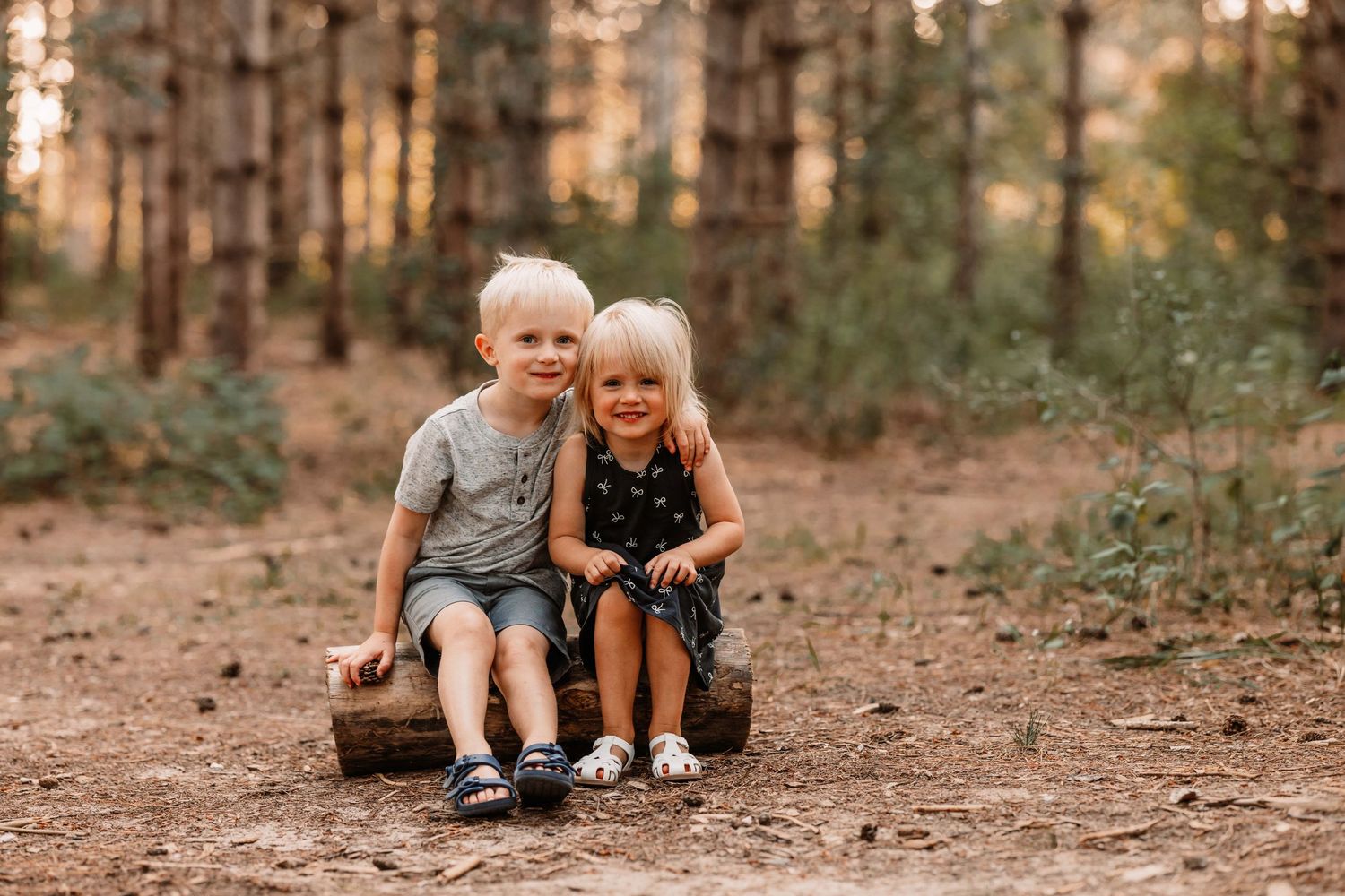 Two children sitting together on log in pine forest during golden hour.