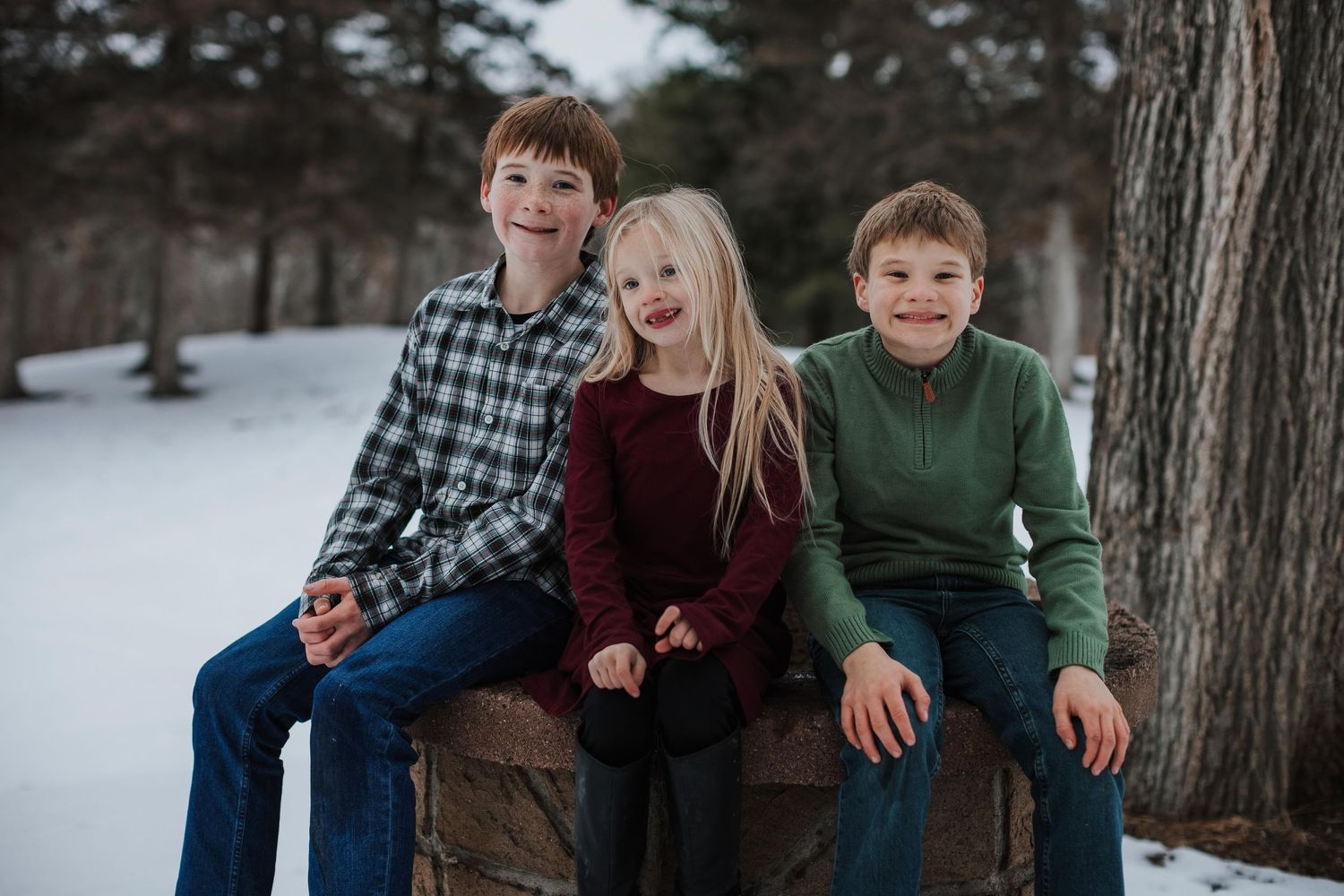 Three children sit together on a wooden bench in a snowy winter setting.