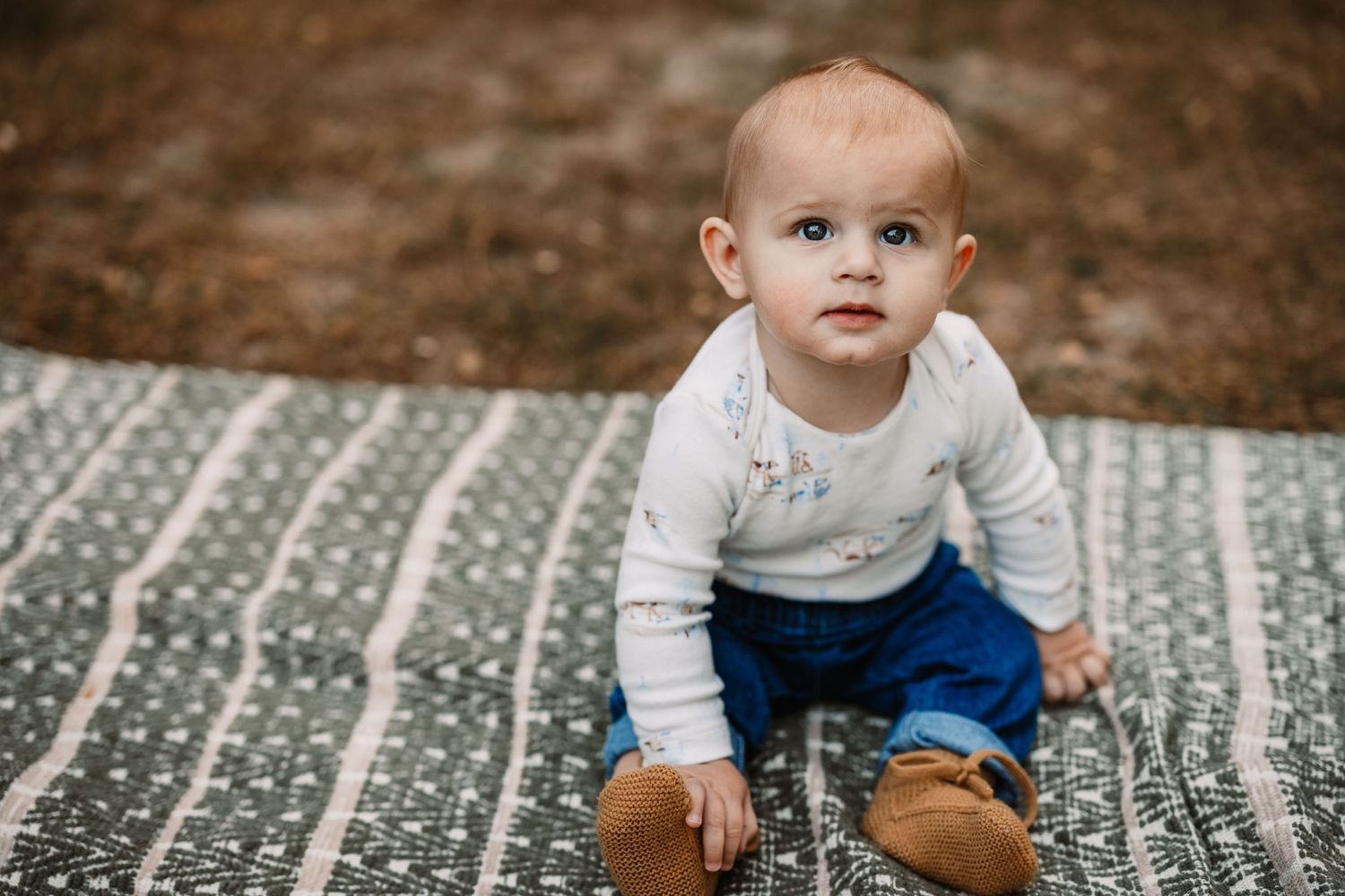 Baby in white sweater and blue pants sitting on patterned blanket outdoors.