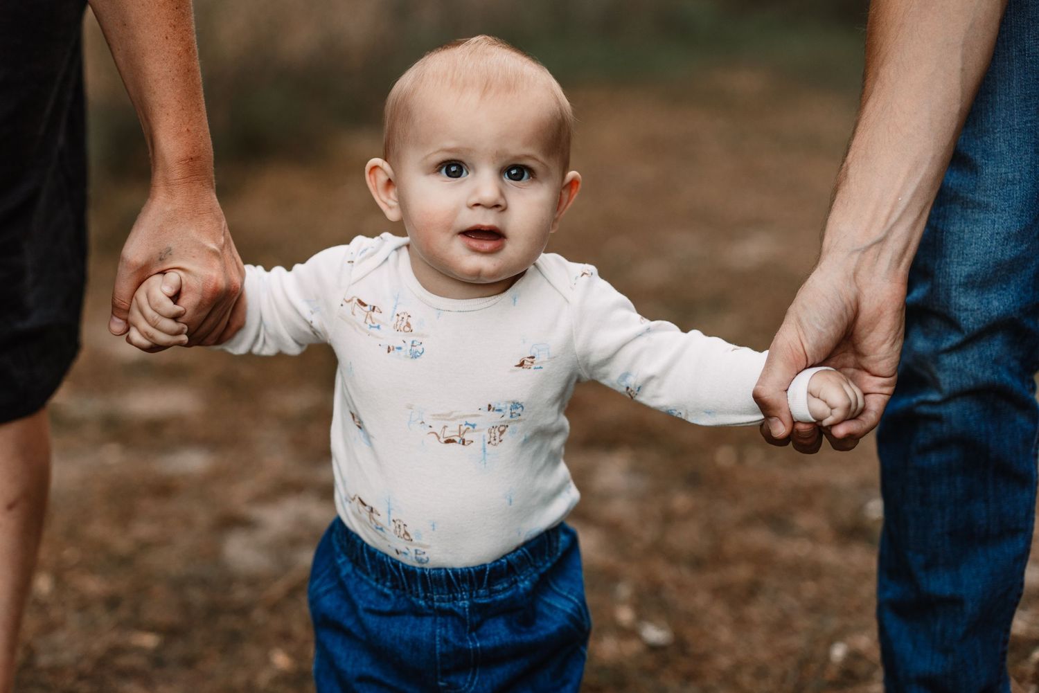 Baby in white top and blue pants learning to walk while holding hands with adults.
