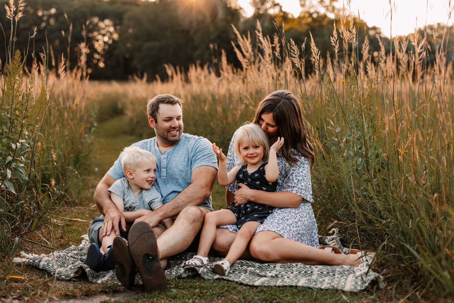 Family sitting together on blanket in tall golden grass during sunset.