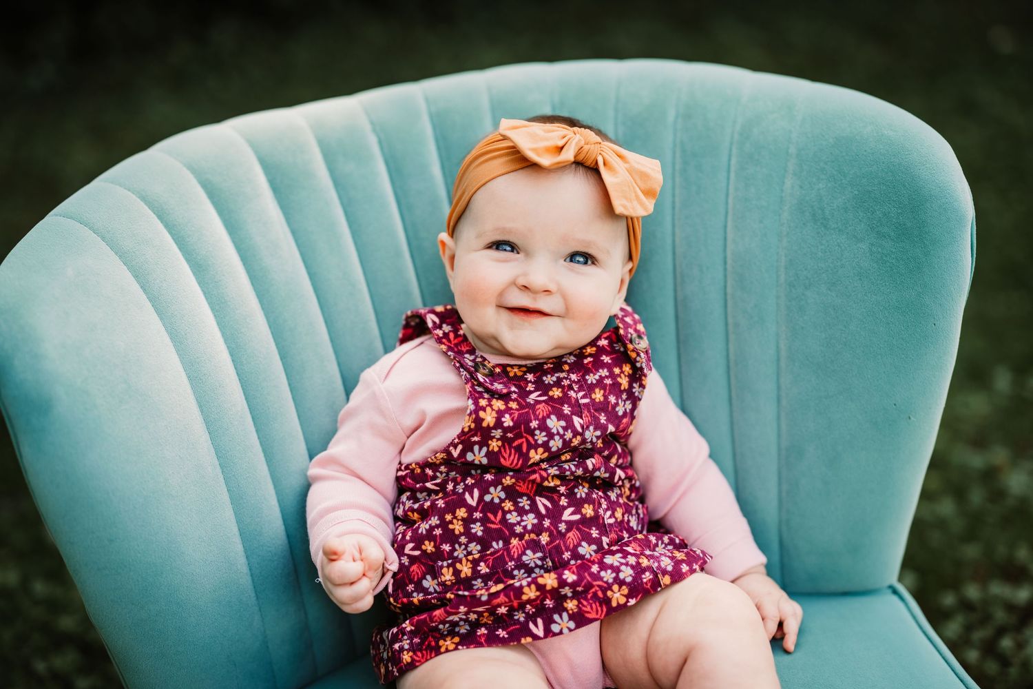 Baby wearing floral dress and bow headband sitting in teal velvet chair.